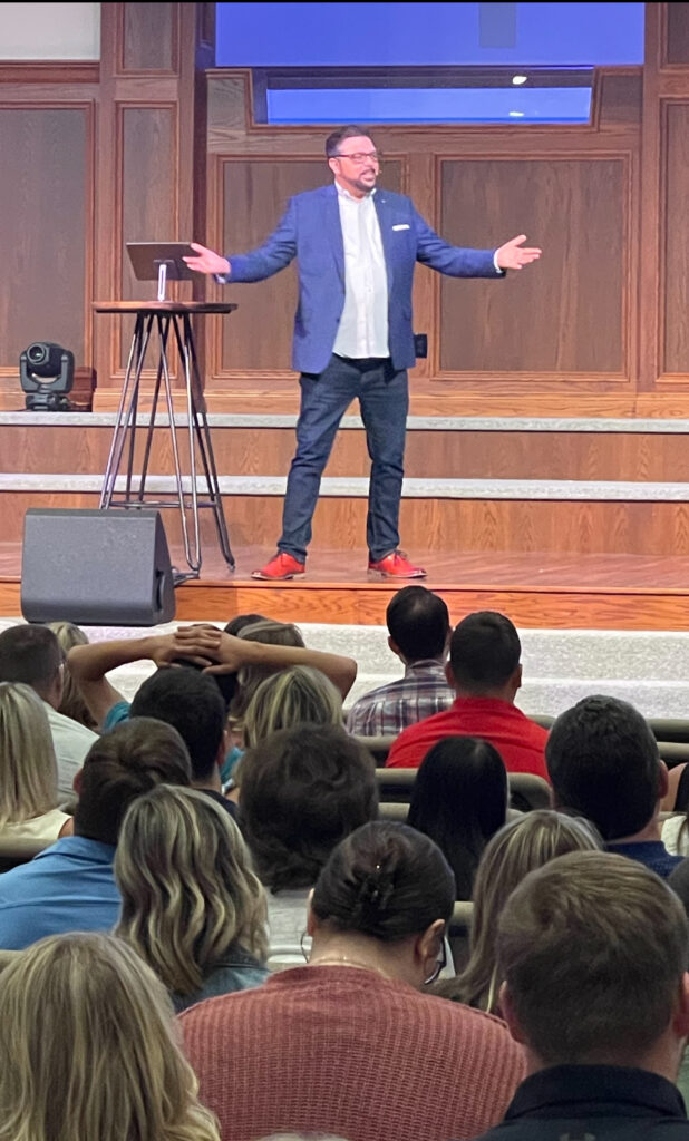 A man in blue jacket standing on stage with arms outstretched.